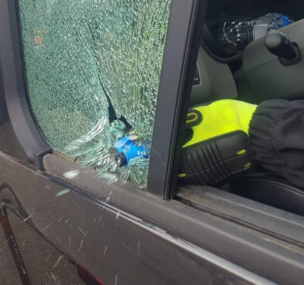 Blaues Rettungswerkzeug an einer zertrümmerten Auto-Seitenscheibe, gehalten mit einem gelb-schwarzen Handschuh.
