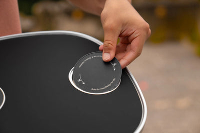 Eine Hand hebt eine runde schwarze Abdeckung aus der schwarzen Platte des Tesla Model 3 Tischs mit silberfarbenem Rand.