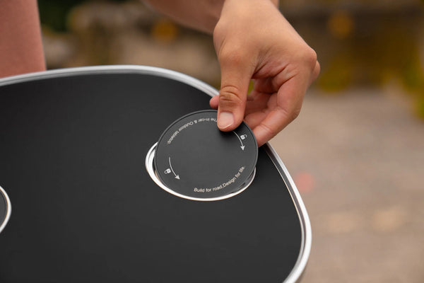 Eine Hand hebt eine runde schwarze Abdeckung aus der schwarzen Platte des Tesla Model 3 Tischs mit silberfarbenem Rand.