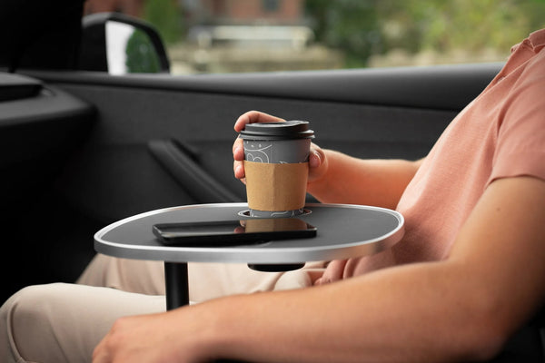 Beifahrer hält einen Kaffeebecher über den Getränkehalter eines schwarzen Tesla Model 3 Tischs; auf der Platte liegt ein Smartphone.