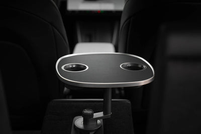 Schwarzer Tesla Model 3 Tisch mit zwei Getränkehaltern und silberfarbenem Rand, von der Rückbank zwischen den Vordersitzen gesehen.