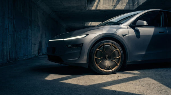 Tesla Model Y Juniper Felgen CHELA in Matte Bronze und Satin Carbon am grauen Fahrzeug, Nahaufnahme vor moderner Betonarchitektur.