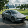 Graues Tesla Model Y Juniper mit CHELA Felgen in Glossy Titanium und Satin Carbon, schräge Frontansicht auf nasser Fahrbahn.