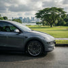Graues Tesla Model Y Juniper mit CHELA 20-Zoll-Felgen in Glossy Titanium und Satin Carbon, Nahaufnahme im grünen Stadtumfeld.
