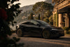 Schwarzes Tesla Model Y Juniper mit CHELA 20-Zoll-Felgen in Matte Bronze und Satin Carbon, Seitenansicht vor einer Villa am Wasser.