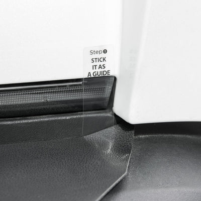 Detail des Tesla Stoßstangenschutzes an einer weißen Karosserie, mit transparenter Ausrichtlasche neben einer Leuchte.